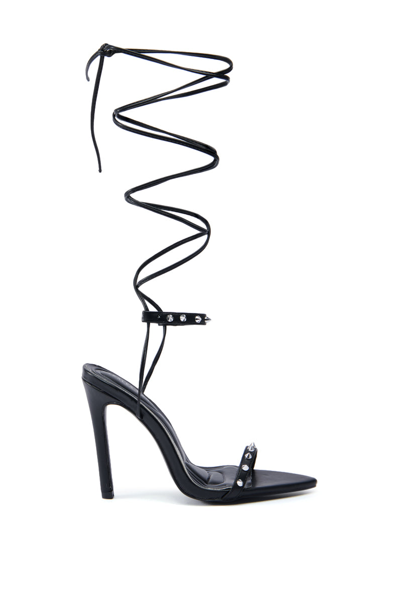The AZALEA WANG JACINTO BLACK SPIKE DETAIL SANDAL features a black high-heeled stiletto with an ankle strap accented by silver studs and a long lace that elegantly wraps around the leg, showcasing a sleek, minimalist design with black spike details against a crisp white background.