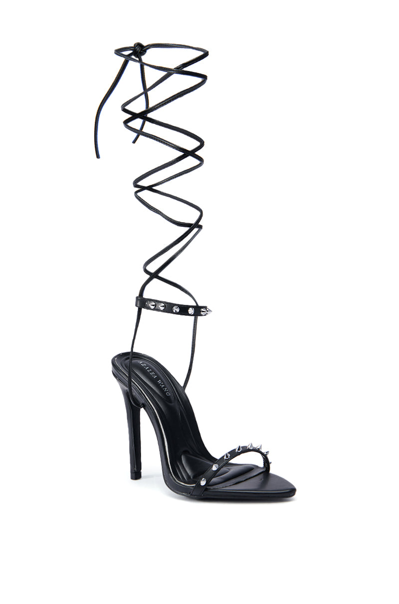 The AZALEA WANG JACINTO BLACK SPIKE DETAIL SANDAL boasts a striking black stiletto heel complemented by an ankle strap embellished with silver spikes. Its extended, slender wraparound lace delicately coils upwards, while the chic open toe design enhances its elegance.