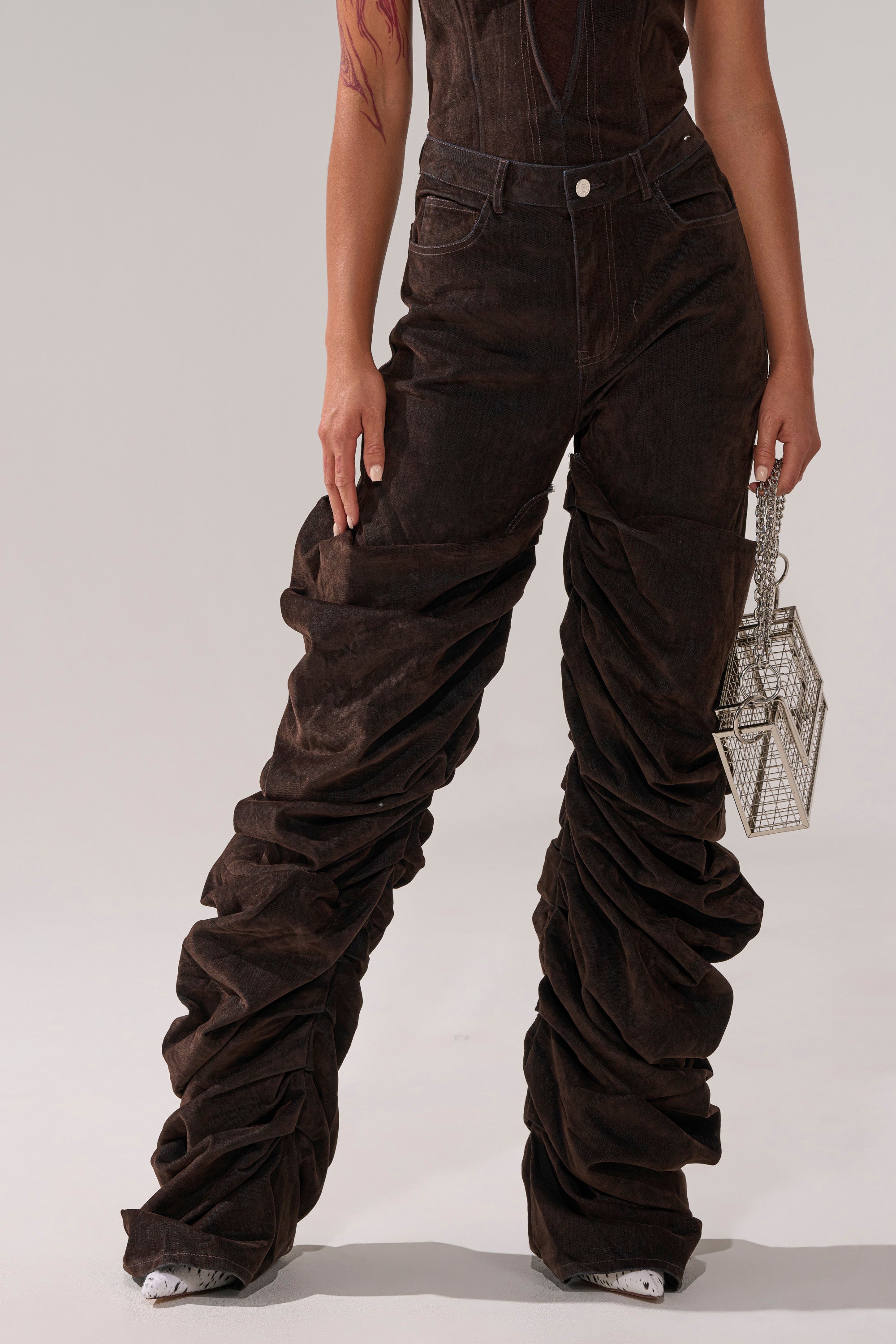 A person models the OH BABY VELVET DENIM PANT—high-rise, relaxed fit, dark brown with ruching—and holds a geometric purse against a plain background. Their face is not shown.