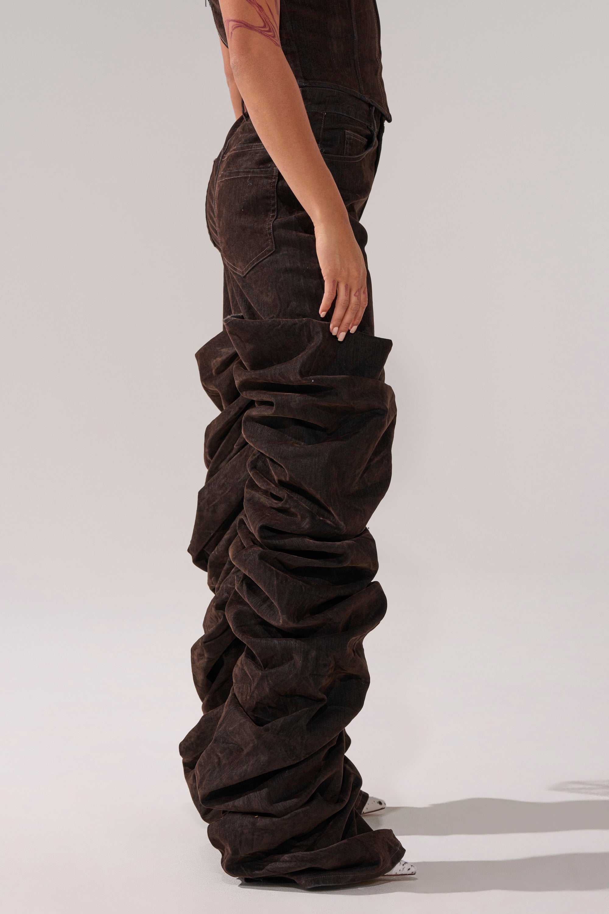 A person in profile wears the OH BABY VELVET DENIM PANT—dark brown, high-rise, relaxed fit, extremely oversized and heavily ruched from ankles to thighs—paired with a brown fitted top.