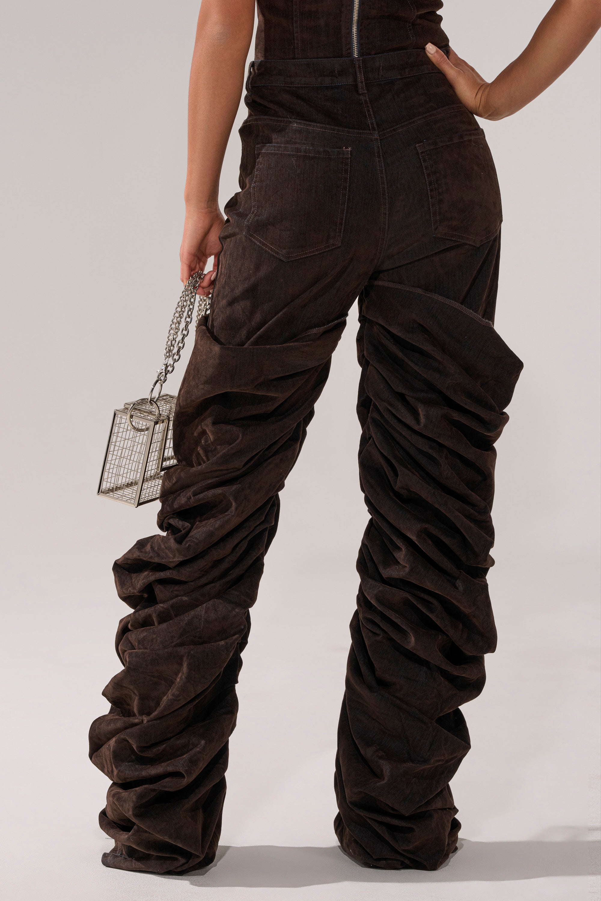 A person stands facing away in OH BABY VELVET DENIM PANT—dark brown, high-rise relaxed fit with dramatic ruched overlay—holding a small metallic box-shaped purse with chain strap. The background is plain and light-colored.