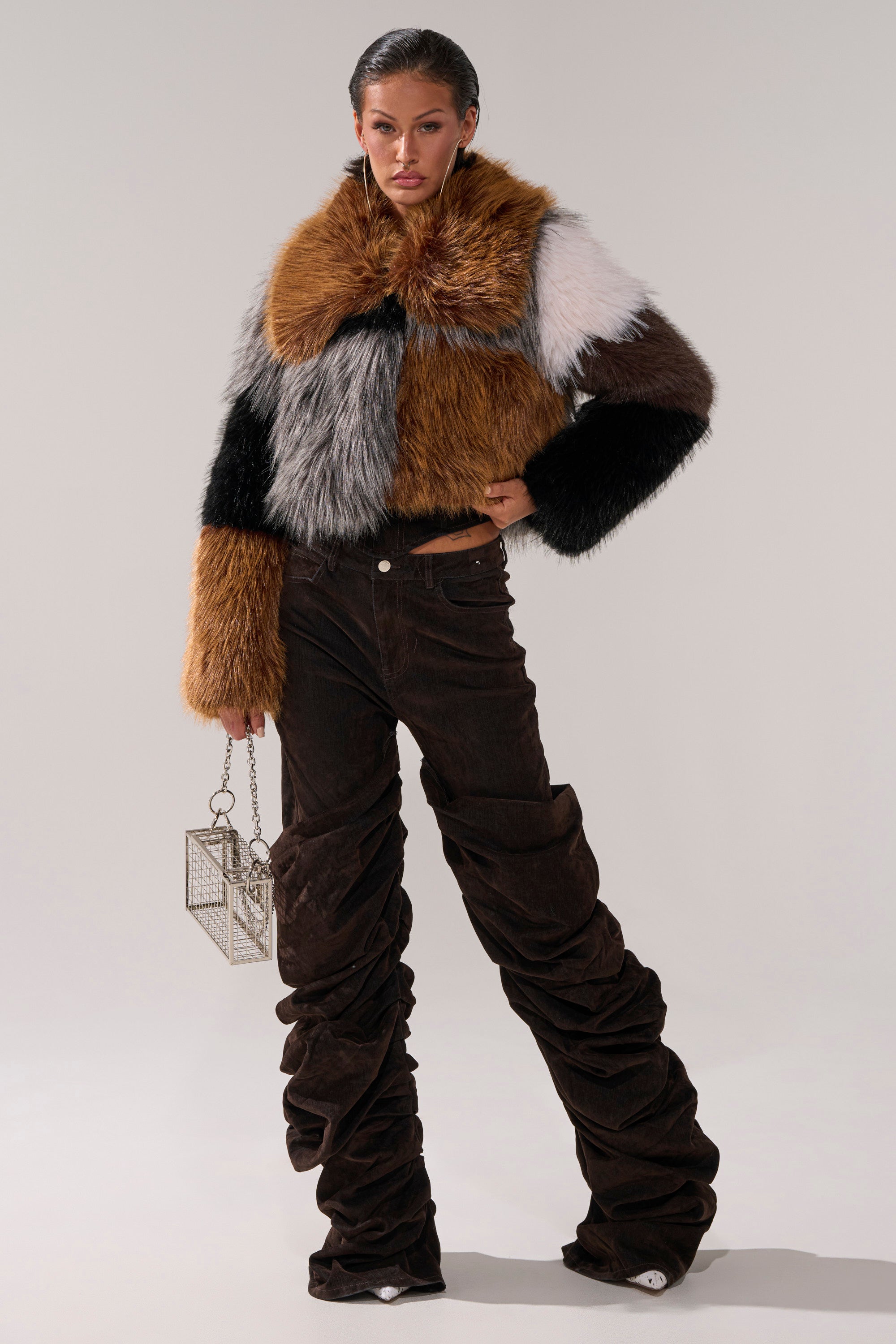 A woman stands against a plain light background wearing the OH BABY VELVET DENIM PANT with a colorful, oversized faux fur jacket and holding a small geometric metallic handbag.