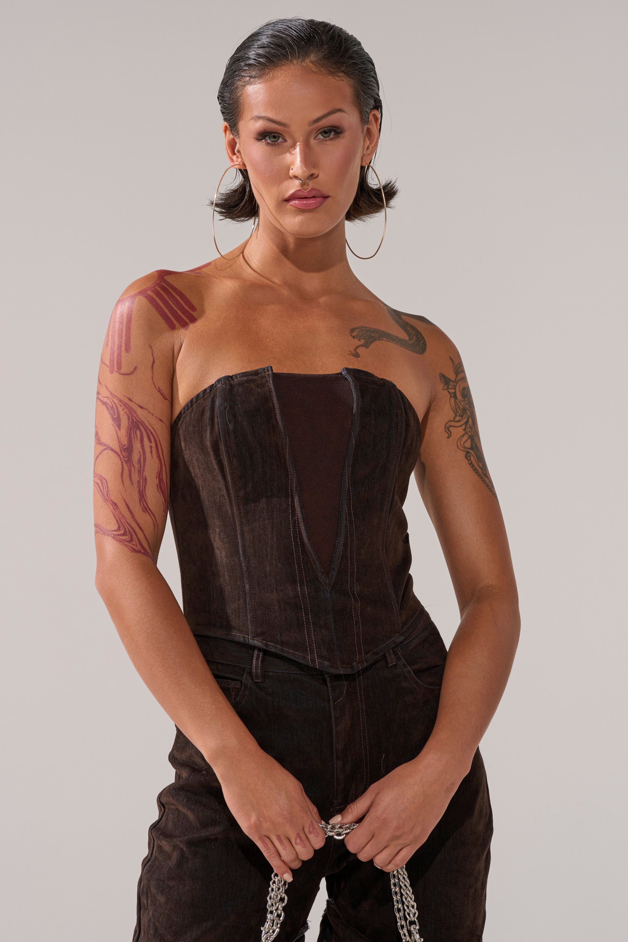A woman with tattoos wears the OH BABY VELVET DENIM CORSET in brown with matching pants, large hoop earrings, and a silver chain. She has slicked-back hair and stands against a plain background.