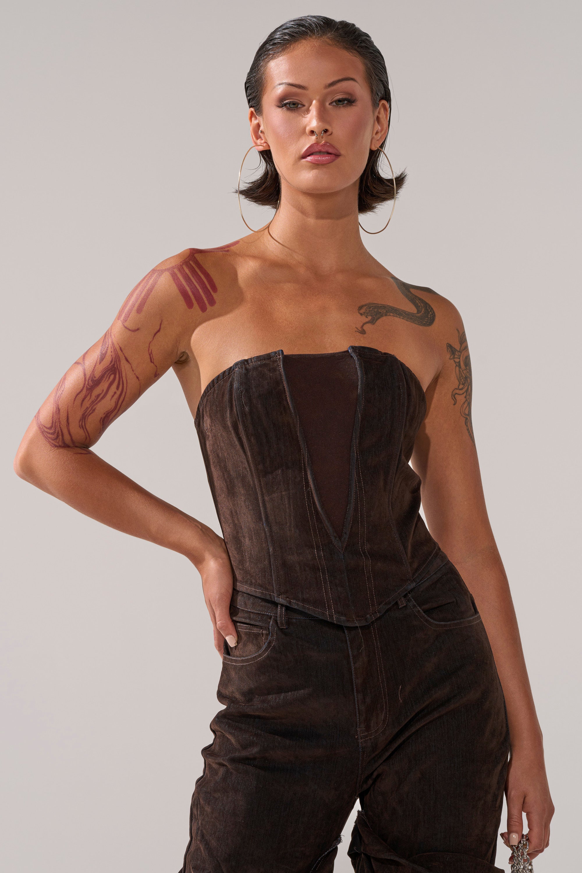 A person with short, slicked-back hair confidently poses against a plain light background, wearing the OH BABY VELVET DENIM CORSET and matching pants that reveal arm and shoulder tattoos.