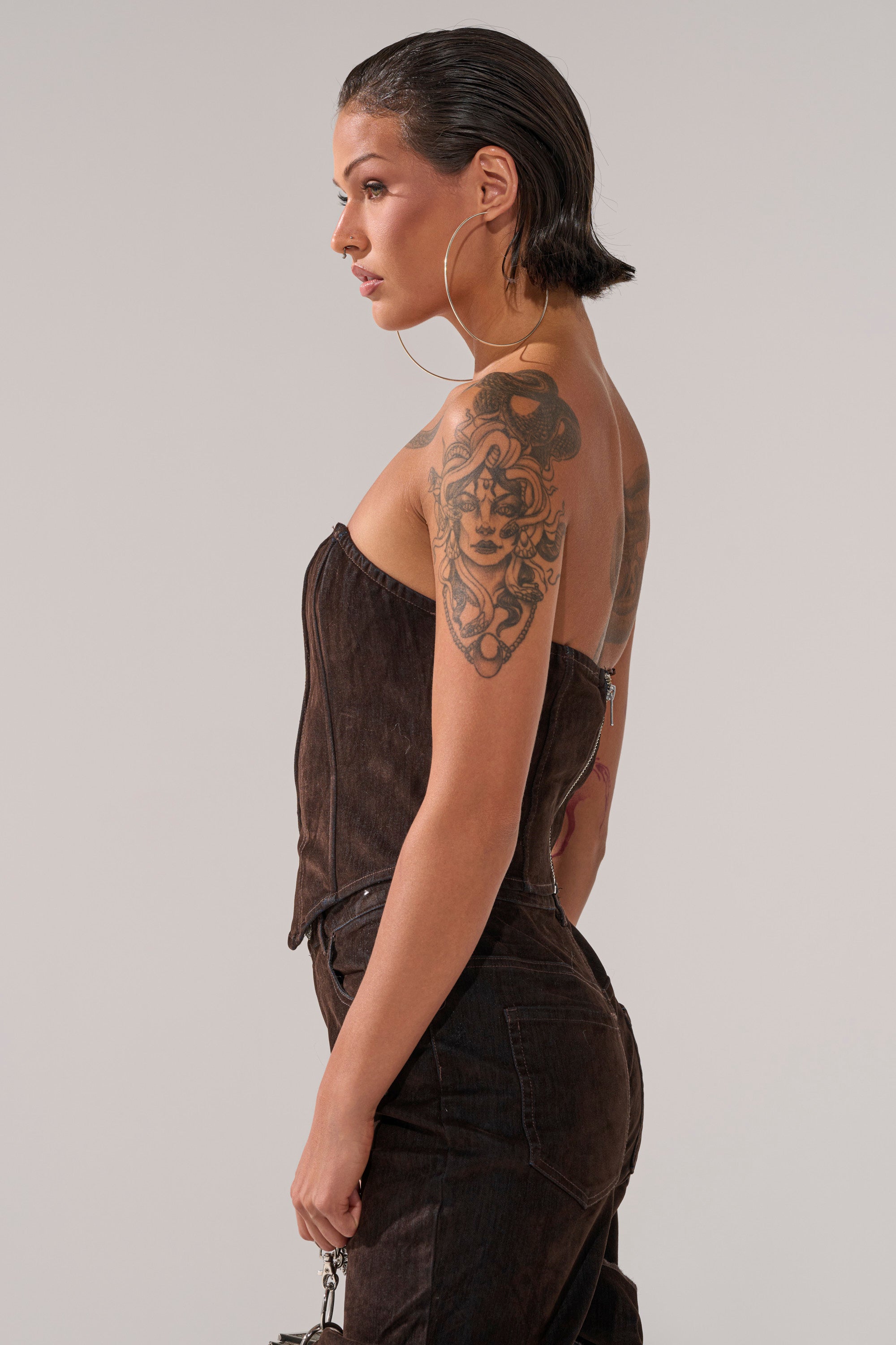A woman with slicked-back dark hair, large hoop earrings, and a detailed upper-arm tattoo stands in profile, wearing the OH BABY VELVET DENIM CORSET with matching pants against a plain background.