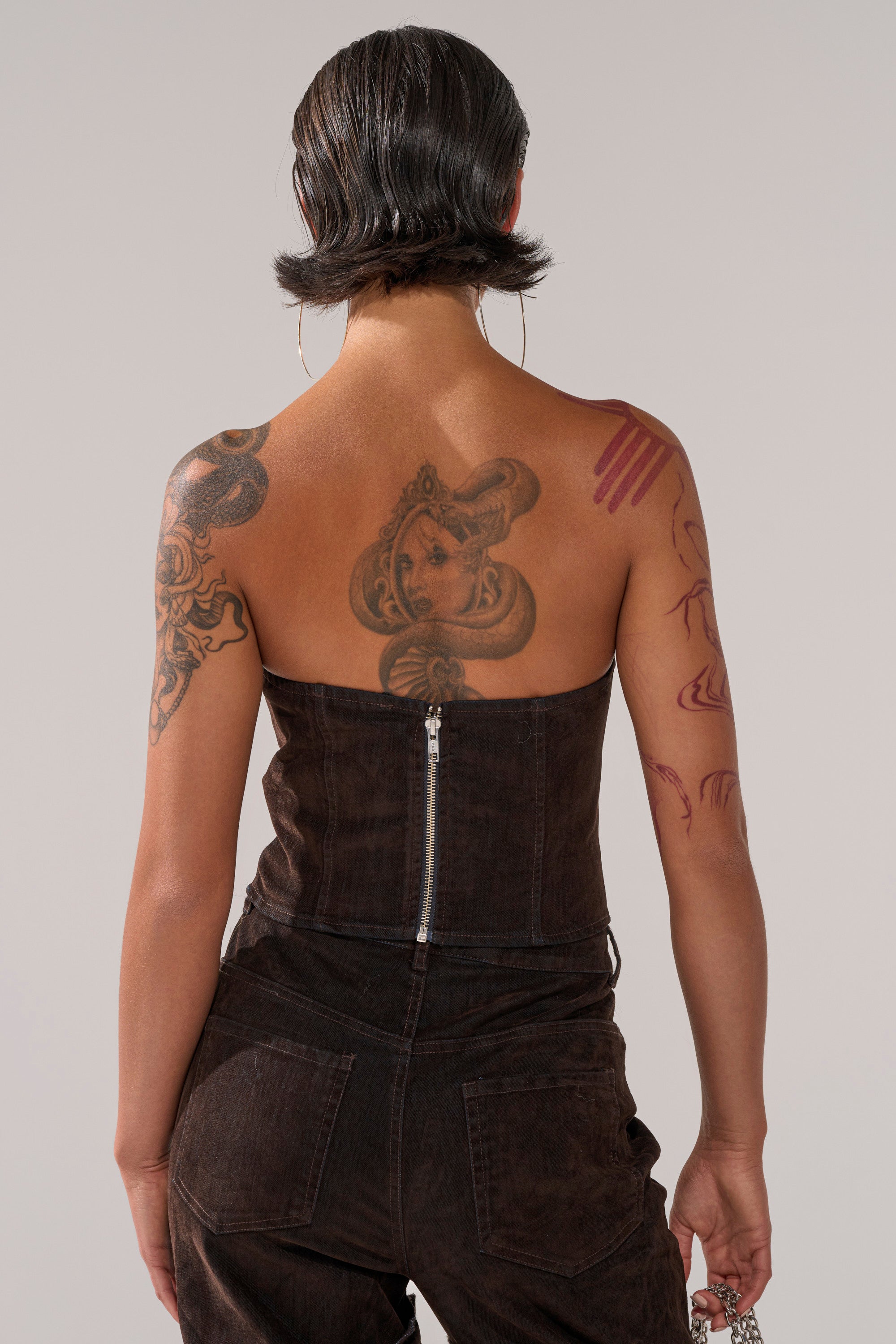 A person with short, dark hair and large back and arm tattoos is shown from behind wearing the OH BABY VELVET DENIM CORSET. They hold a chunky chain in their right hand.