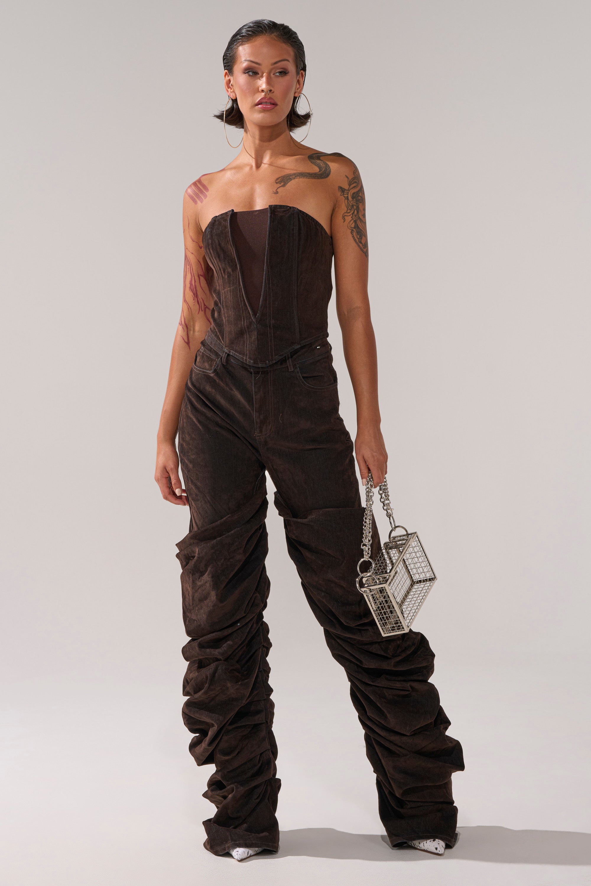 A woman stands confidently in the OH BABY VELVET DENIM CORSET, a strapless brown jumpsuit with exaggerated ruched pant legs, holding a small silver box-shaped handbag. Tattoos are visible on her right arm and chest.