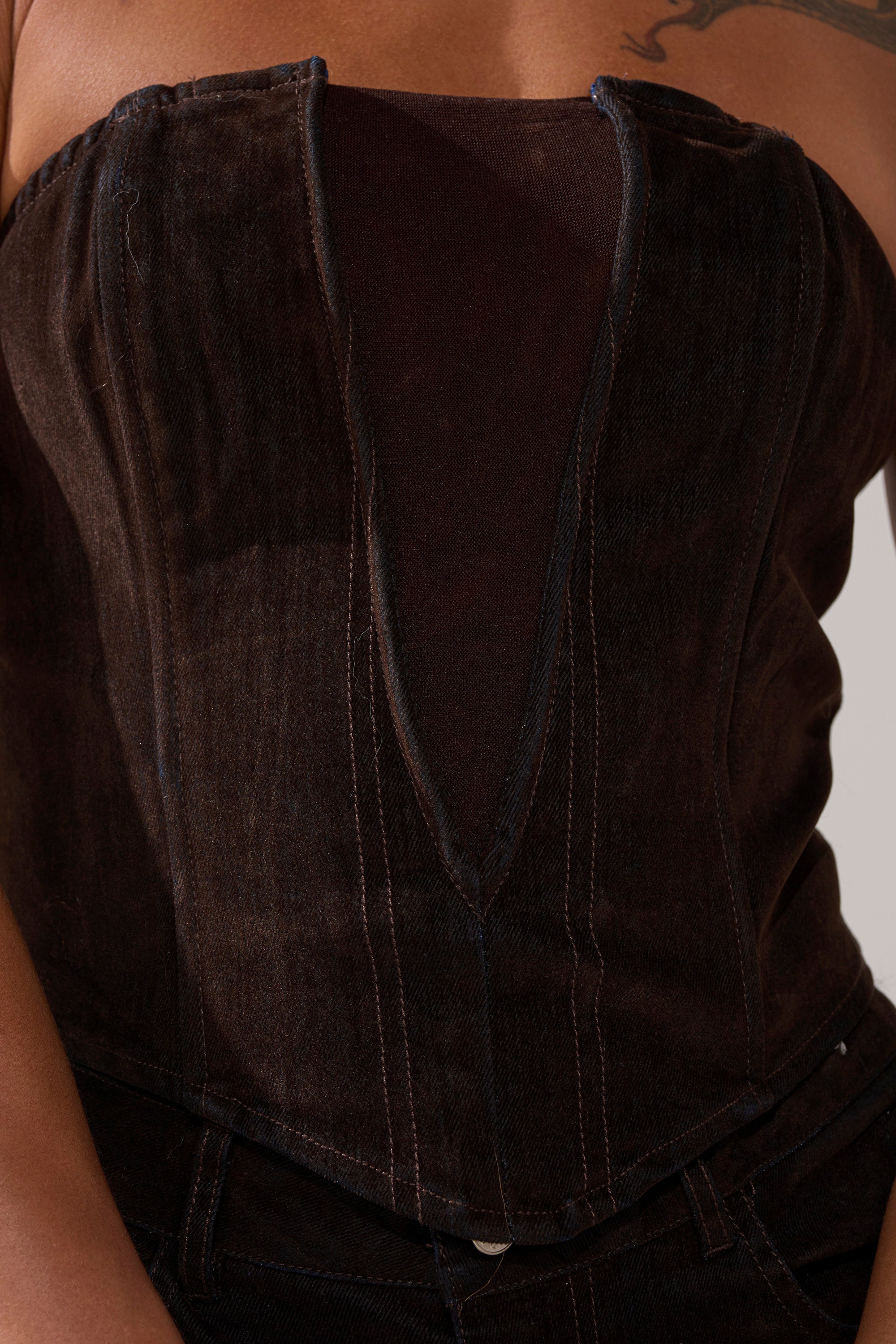 A close-up of someone wearing the OH BABY VELVET DENIM CORSET—a dark brown, strapless top with a deep V-neckline and visible stitching. Part of their arm and a tattoo can also be seen.