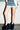 Wearing a frayed blue denim mini skirt, white shirt, bracelets, and AZALEA WANG JAEKE NAVY FUR WEDGE PUMP slippers, a person stands against a white brick wall on a concrete floor.