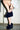 Wearing a white shirt and several bracelets, a person bends to adjust the AZALEA WANG JAEKE NAVY FUR WEDGE PUMP on their feet, standing on a worn light brown floor with a white brick wall in the background.