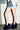 A person wearing a frayed denim mini skirt, white shirt, bracelets, and AZALEA WANG JAEKE NAVY FUR WEDGE PUMP stands against a white wall with exposed pipes. Only their lower body is visible.