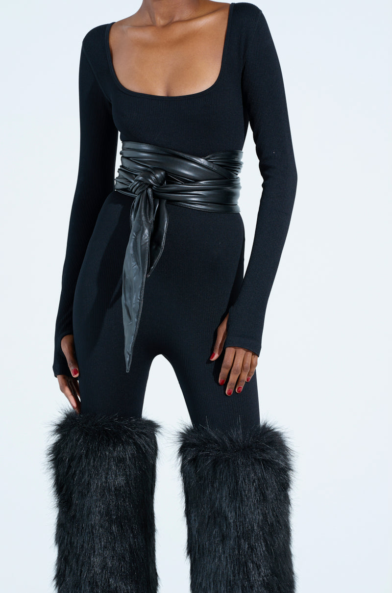 Dressed in a black long-sleeve, scoop-neck outfit paired with the ESSENTIAL WRAP AROUND TIE BLACK BELT, they wear knee-high boots trimmed with faux fur. A sleek faux leather accessory completes the look against a plain white background.