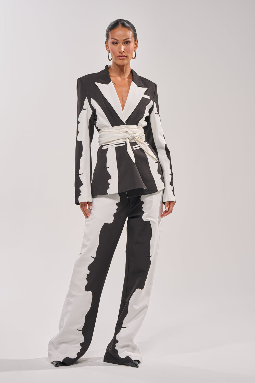 A woman poses confidently against a plain light background, wearing a bold black and white pantsuit with abstract face designs, styled with the ULTIMATE CINCH PU BELT—a white faux leather bow belt that stands out as a statement accessory. 1