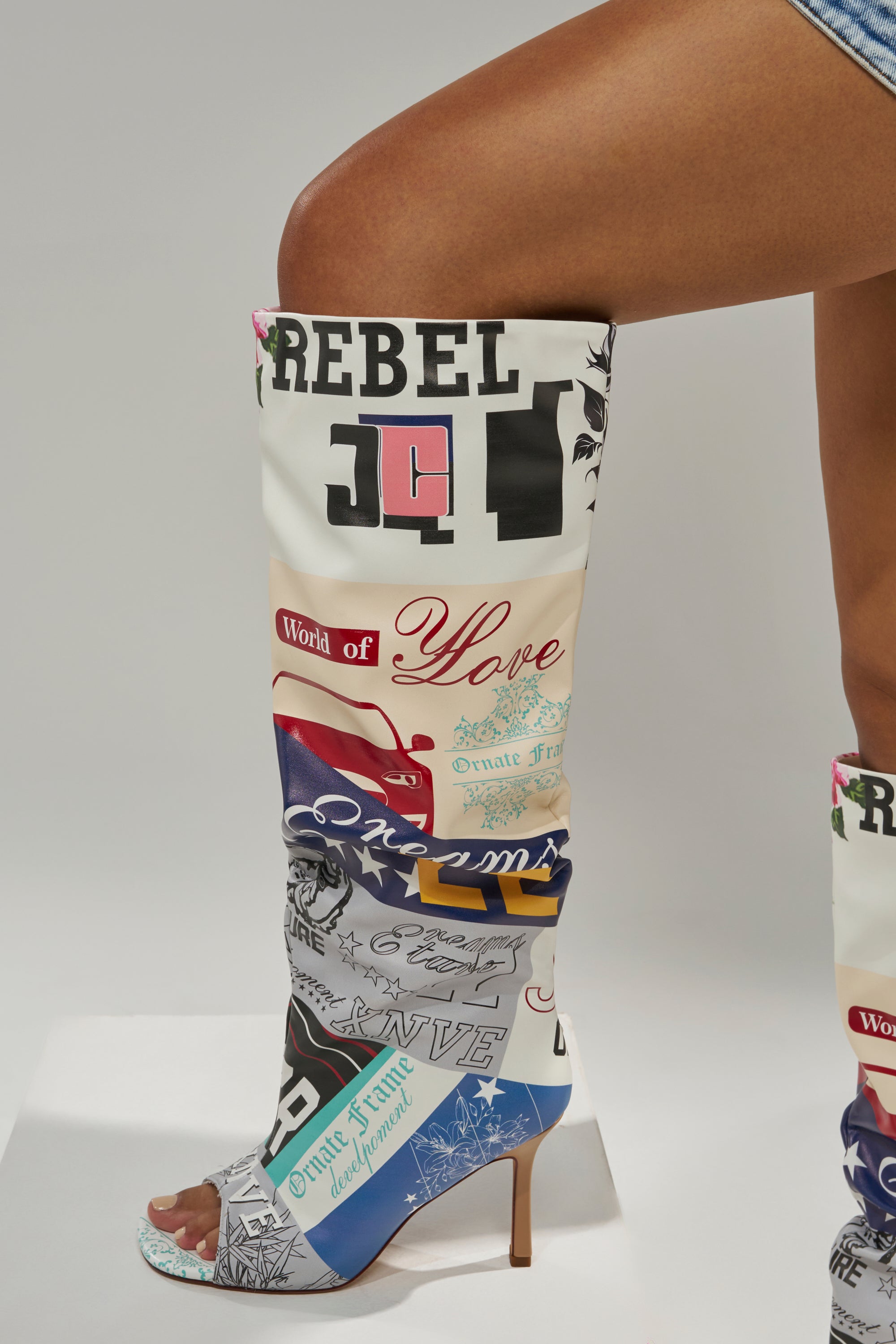 A person wears the AZALEA WANG JALISA MULTI NOVELTY PRINTED OPEN TOE BOOT, featuring knee-high patchwork with graffiti-style words like “REBEL” and “Love,” paired with denim shorts against a plain white background.