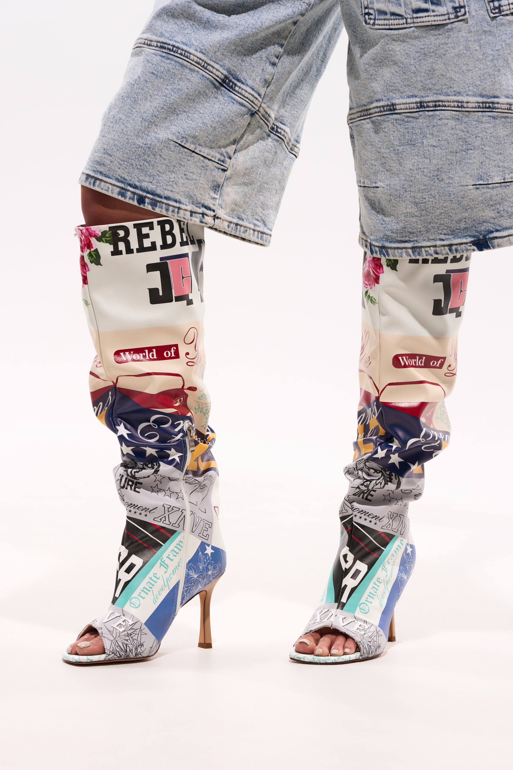 Someone wears rolled-up, light-wash denim shorts paired with the AZALEA WANG JALISA MULTI NOVELTY PRINTED OPEN TOE BOOT, featuring graphic, text, and floral patchwork designs on a white background. 0