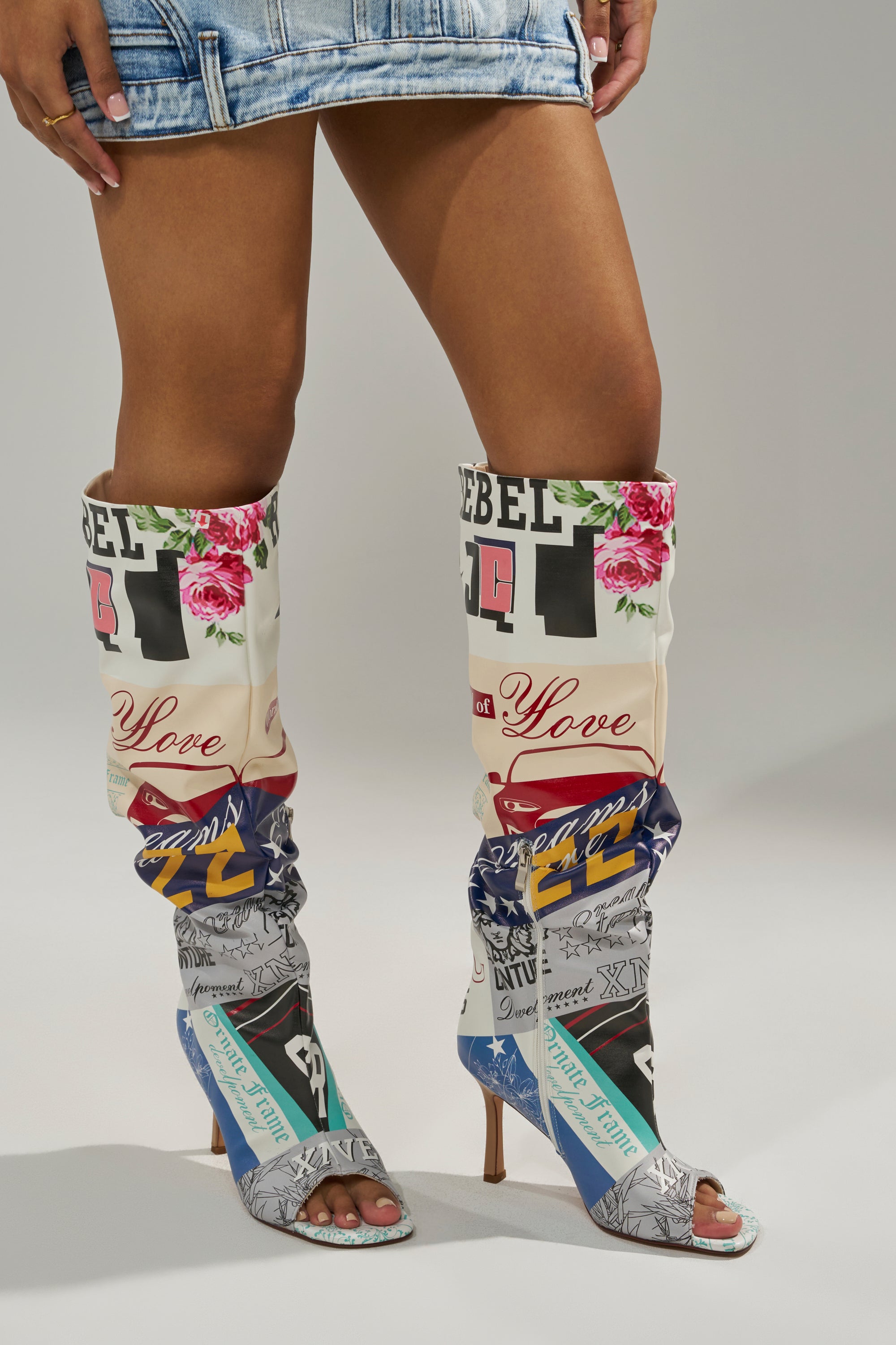 Someone wears a short denim skirt and AZALEA WANG JALISA MULTI NOVELTY PRINTED OPEN TOE BOOTS, featuring vibrant patchwork designs with various prints and text, while standing against a plain gray background.