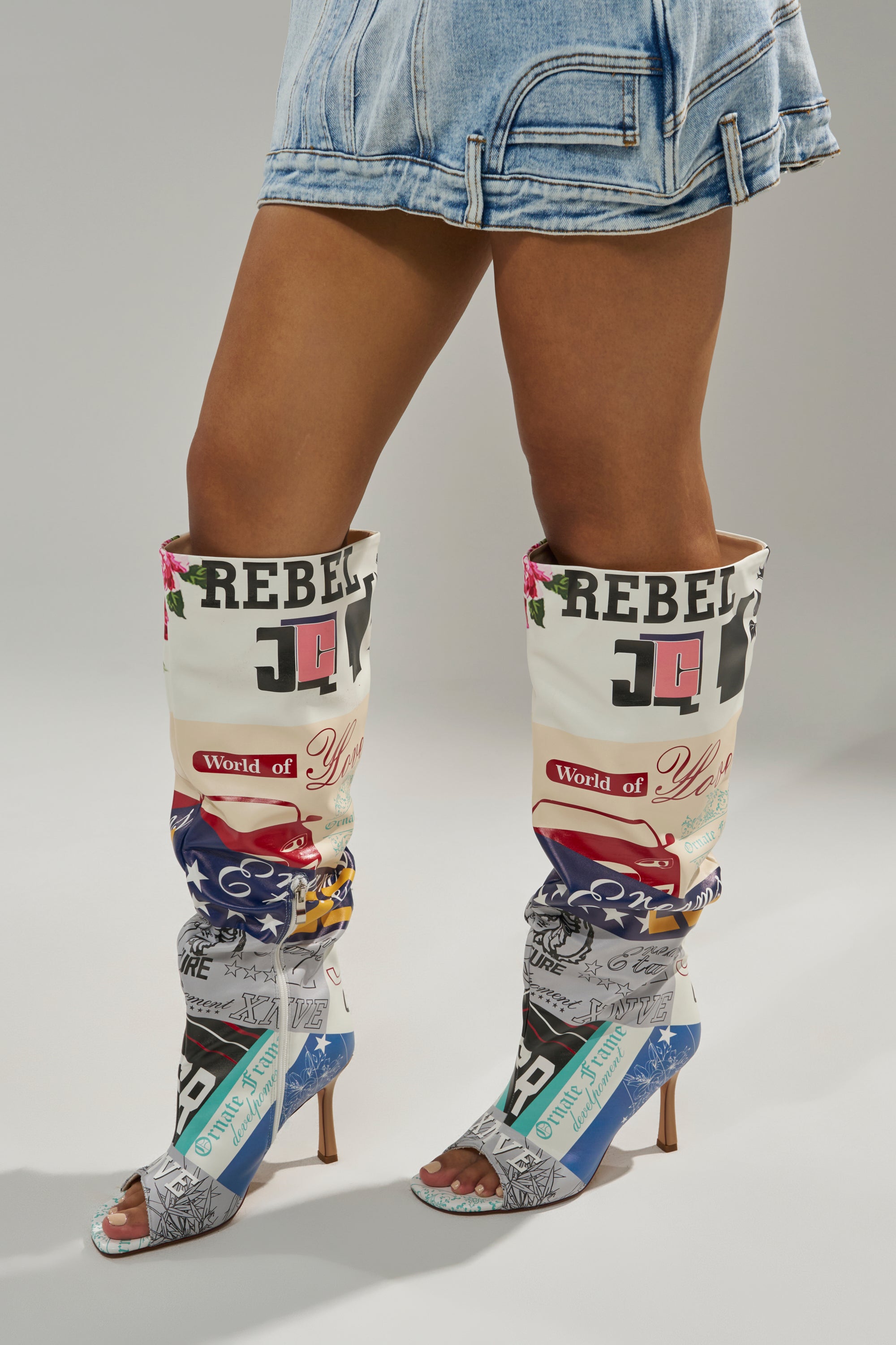 A person wears a short denim skirt and the AZALEA WANG JALISA MULTI NOVELTY PRINTED OPEN TOE BOOT, featuring bold patchwork graphics and phrases like "REBEL" and "World of," standing out against a simple light background.
