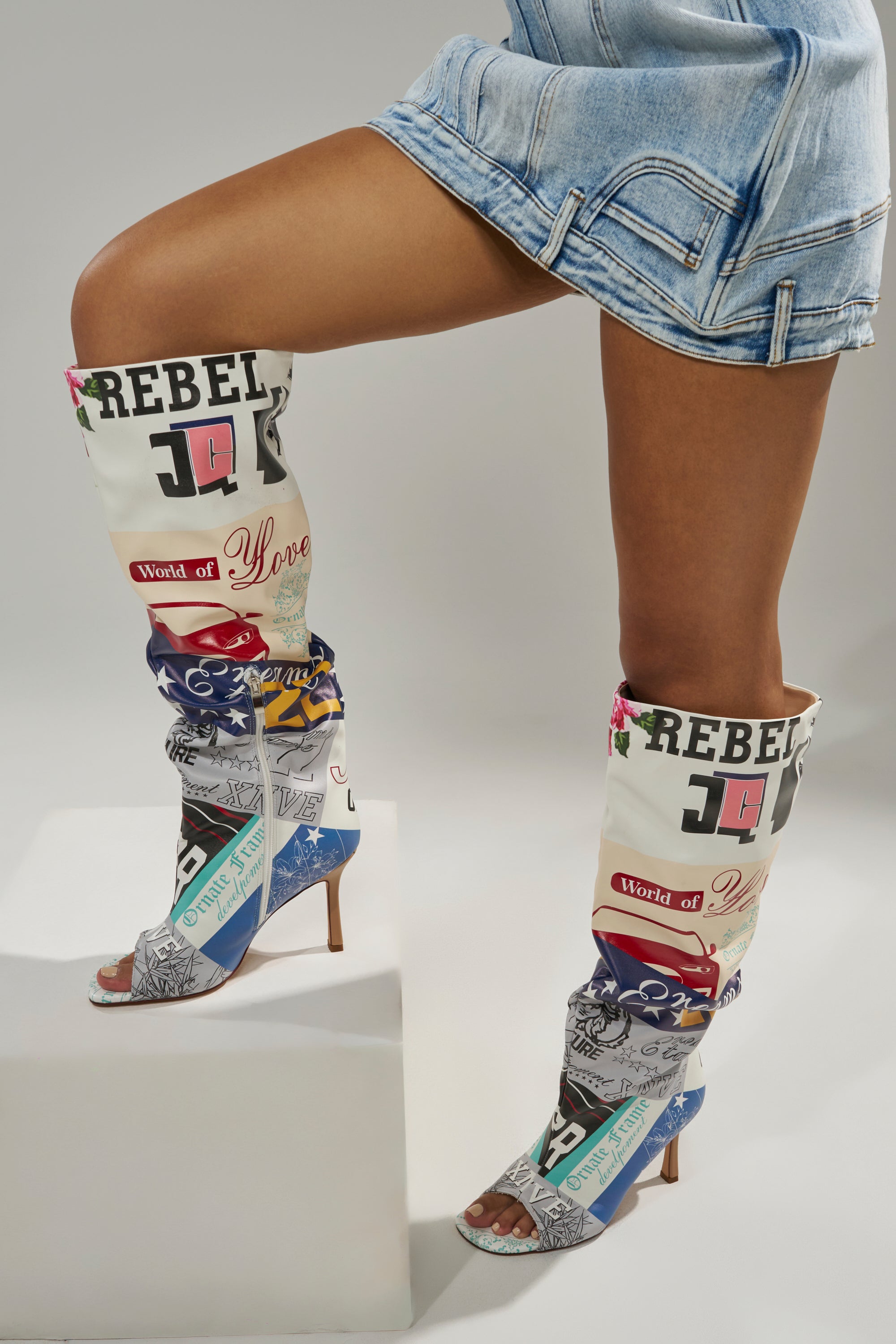A person wears a light denim mini skirt and the AZALEA WANG JALISA MULTI NOVELTY PRINTED OPEN TOE BOOT—colorful knee-high boots with bold graphics and text—standing with one leg bent, foot resting on a white block.