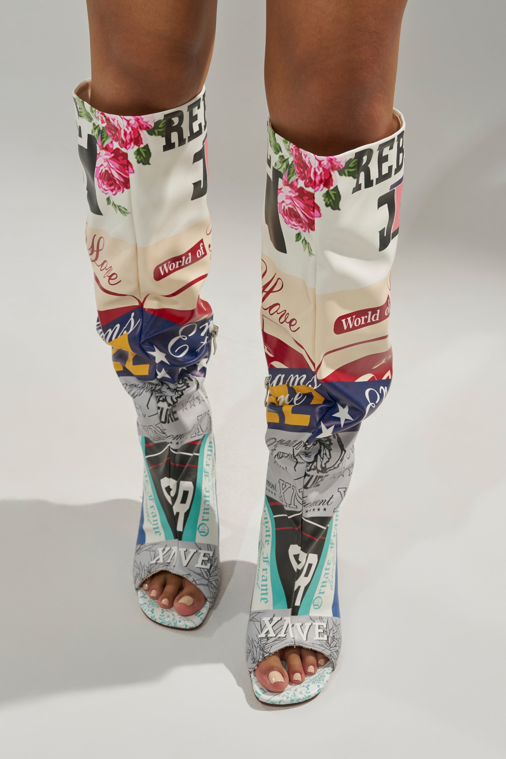 Wearing the AZALEA WANG JALISA MULTI NOVELTY PRINTED OPEN TOE BOOT, which features colorful patterns, bold text, florals, American flag motifs, and graphics on a light background. The knee-high boots are stiletto-heeled and slightly slouched at the calves.