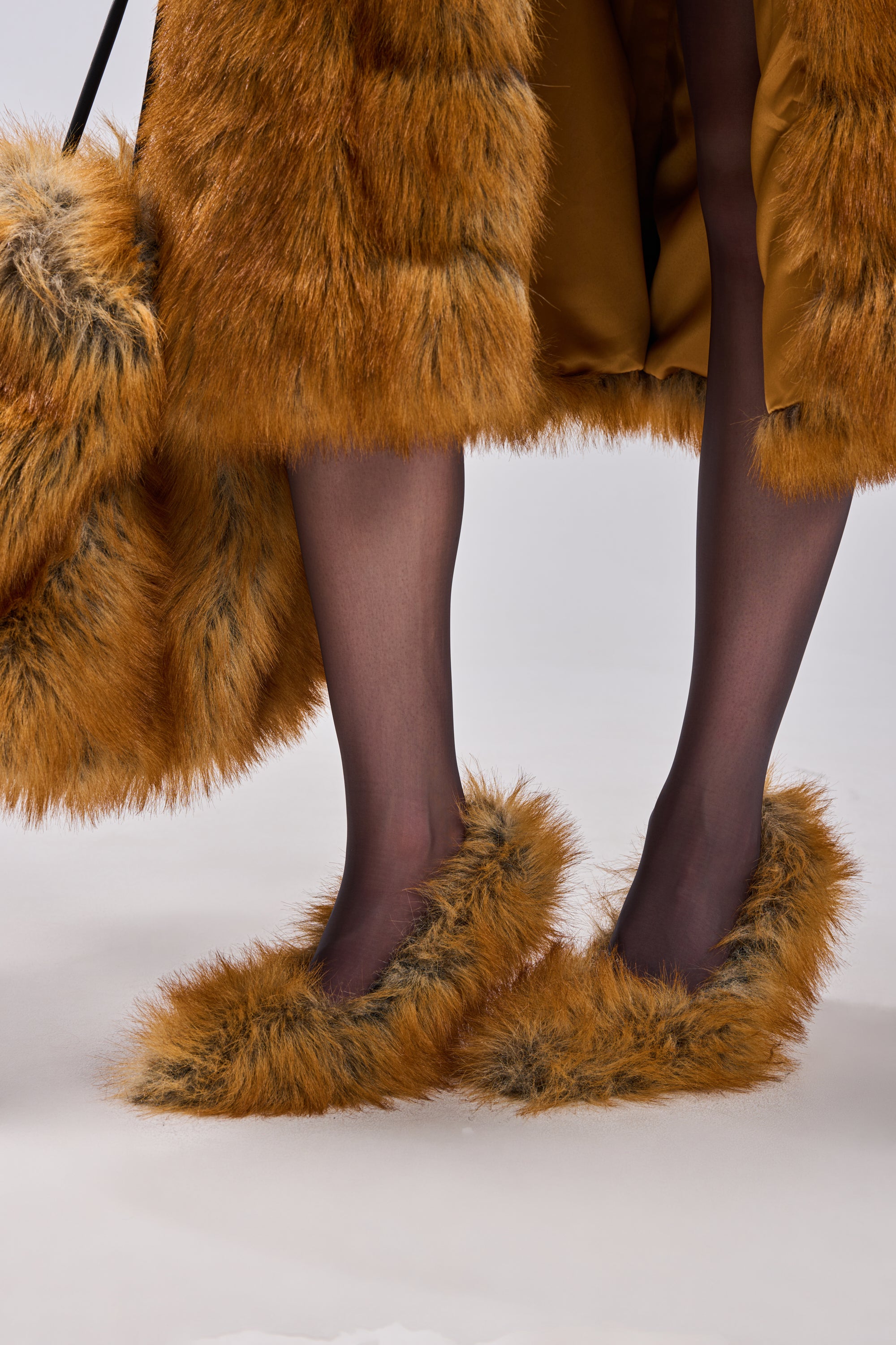 A person wearing black sheer tights and AZALEA WANG JAMEECK TAN FUR PUMP heels, paired with a matching orange faux fur coat and fluffy orange faux fur bag. The image highlights the lower legs and feet.