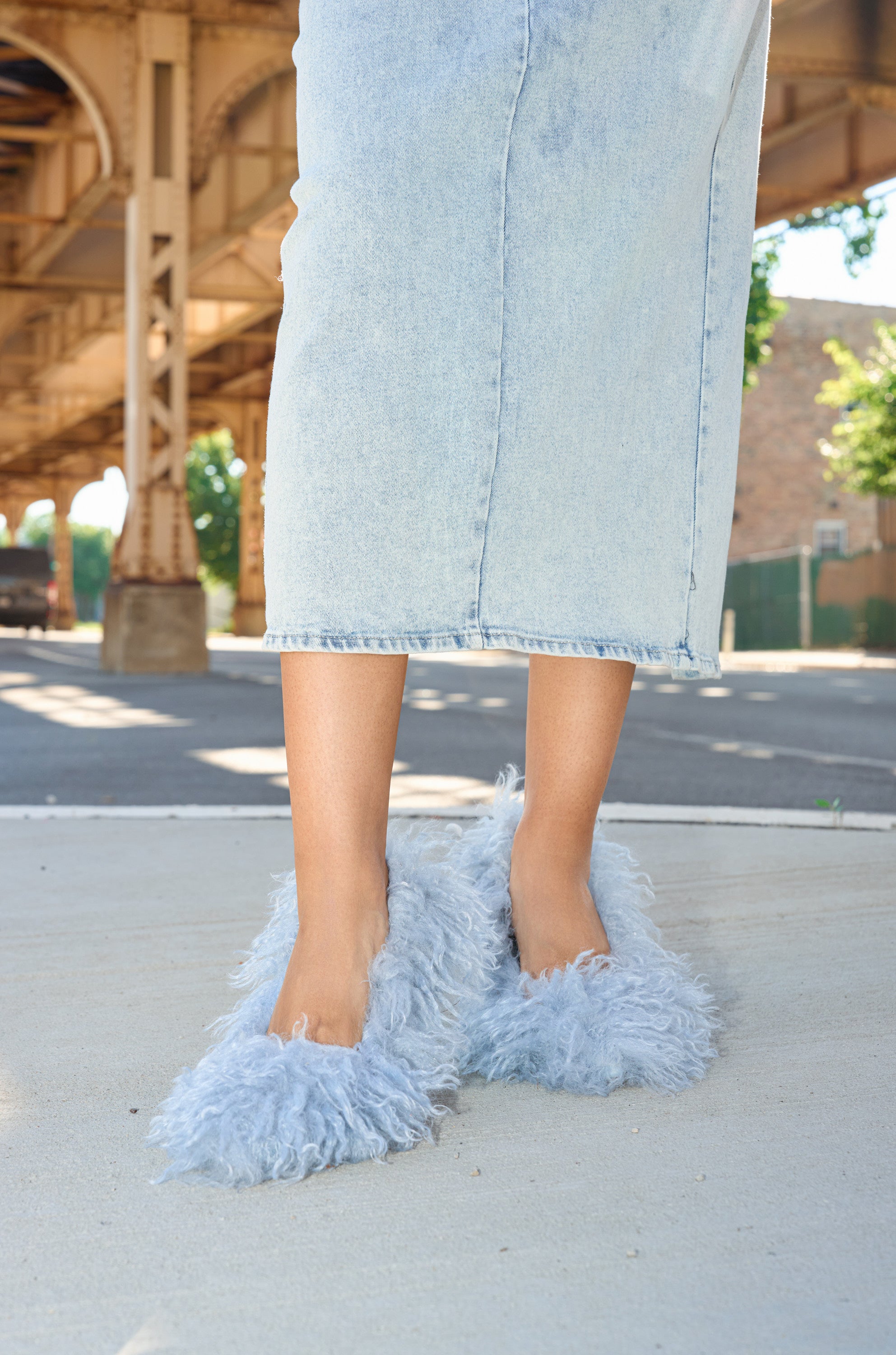 A person stands outdoors on a sidewalk under an elevated train track, wearing cropped light-wash denim jeans and the AZALEA WANG JAMEEKA BABY BLUE CURLY FUR PUMP STILETTO HEEL.