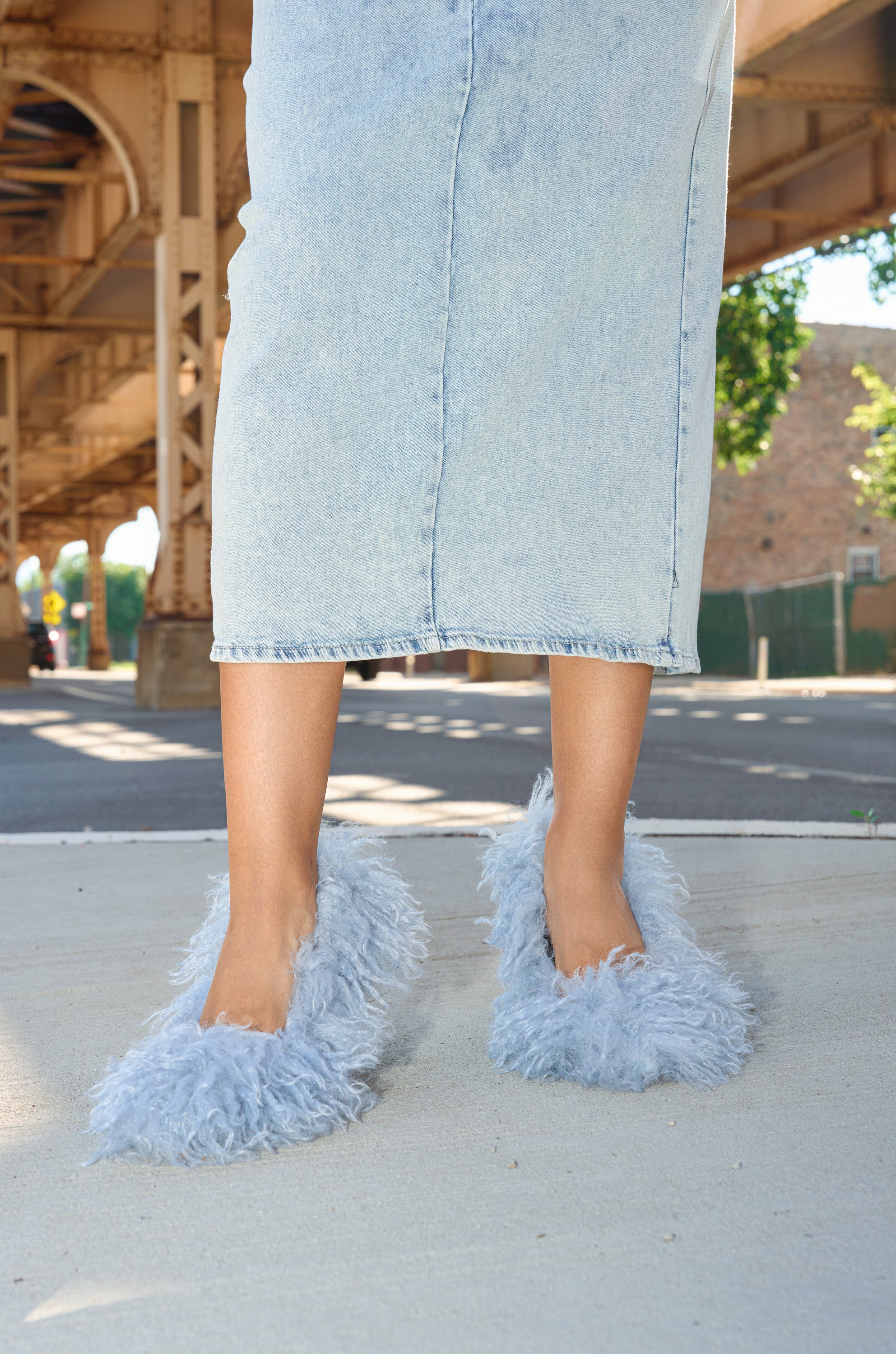 Wearing a light denim skirt and AZALEA WANG JAMEEKA BABY BLUE CURLY FUR PUMP STILETTO HEEL, a person stands on a sidewalk under an overpass with sunlight and trees in the background.