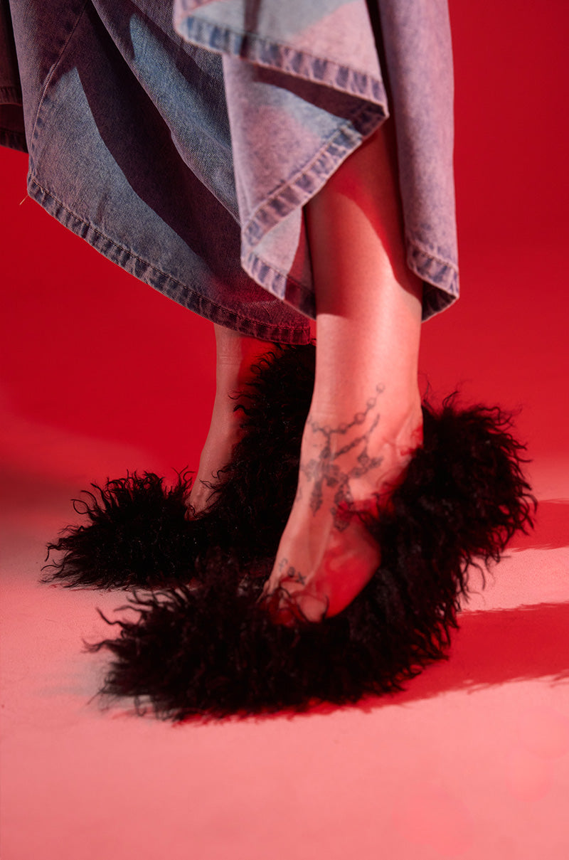 A person stands on a red-lit floor, wearing a long denim skirt and the AZALEA WANG JAMEELA BLACK FUR STILETTO PUMP, with a tattoo visible on their foot.