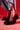A person stands on a red-lit floor, wearing a long denim skirt and the AZALEA WANG JAMEELA BLACK FUR STILETTO PUMP, with a tattoo visible on their foot.