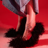 A person stands on a red-lit floor, wearing a long denim skirt and the AZALEA WANG JAMEELA BLACK FUR STILETTO PUMP, with a tattoo visible on their foot.