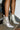 A person wearing the AZALEA WANG JAMILA DRIPPING DIAMONDS SILVER FRINGE BOOTIE, featuring a rhinestone-embellished heel, stands on a metal staircase against a white brick wall backdrop.