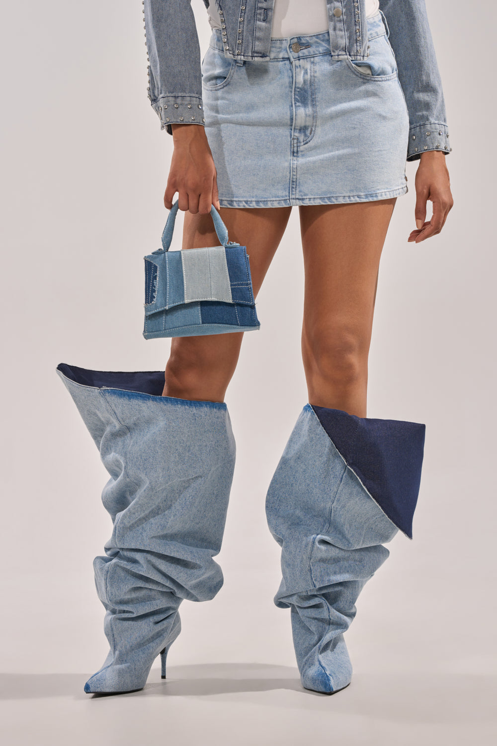 A person stands against a plain background wearing an acid-wash denim miniskirt, matching jacket, and AKIRA GOLD JANELISSE DENIM STILETTO BOOT, holding a small patchwork denim handbag. 2