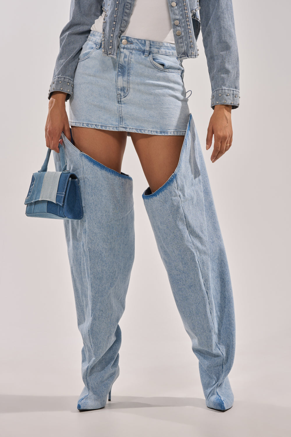 A person wears a light blue denim mini skirt, matching cropped jacket, and AKIRA GOLD JANELISSE DENIM STILETTO BOOTS—unique thigh-high boots with pointed toes resembling pant legs—while holding a small blue handbag. 7