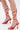 Close-up of a person wearing the AKIRA JANIKA RED HARDWARE STRAPPY SANDAL, featuring red stiletto heels with thin straps wrapping around the ankles and lower legs, tied in bows at the back, against a plain white background.