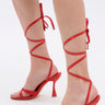 Close-up of a person wearing the AKIRA JANIKA RED HARDWARE STRAPPY SANDAL, featuring red stiletto heels with thin straps wrapping around the ankles and lower legs, tied in bows at the back, against a plain white background.