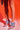Against a red background, a person shows off their legs from mid-thigh down while wearing the AZALEA WANG JANSEN BLUE SNAKE STILETTO SANDAL with a chic snakeskin pattern and holding a sparkly handbag.