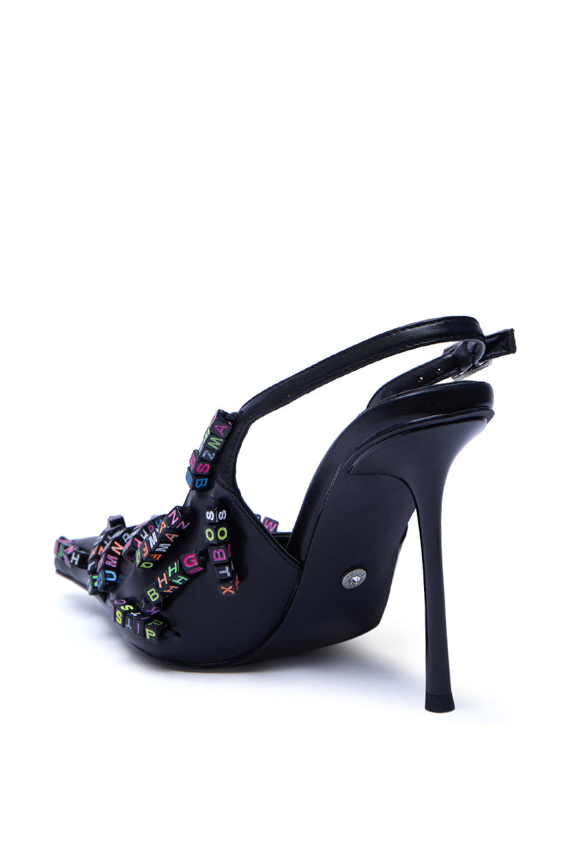 The AKIRA LETTER BEAD EMBELLISHED SLING BACK NOVELTY PUMP is a black faux leather stiletto viewed from the side and slightly behind, featuring colorful alphabet beads on the upper straps.