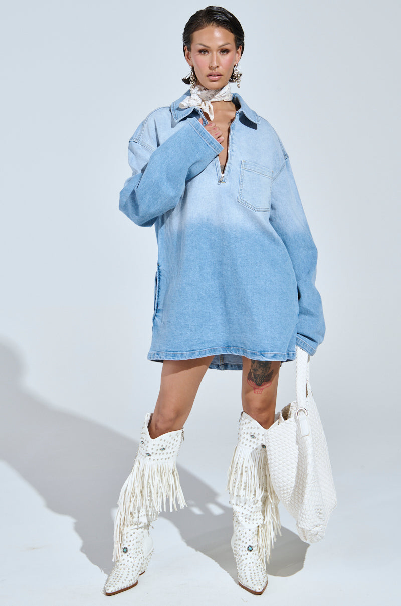 A person confidently poses in a COOL GIRL OVERSIZED OMBRE DENIM SHIRT DRESS with white, fringed knee-high boots. They hold a large white bag, accessorized with statement earrings and a necktie, portraying a casual-cute vibe against a plain light background.