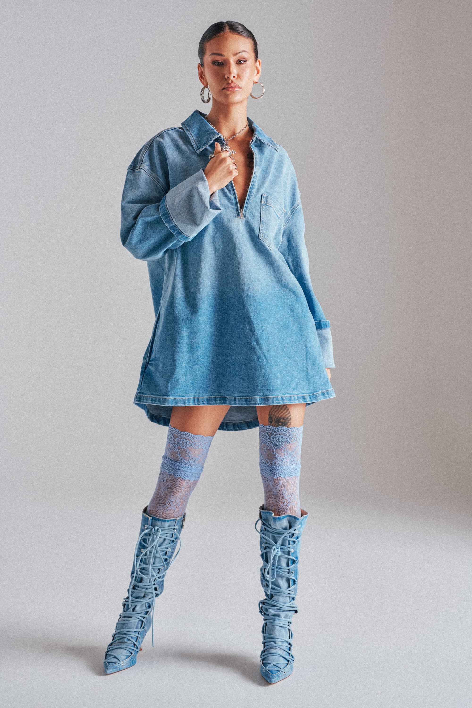 A woman stands confidently in the COOL GIRL OVERSIZED OMBRE DENIM SHIRT DRESS, styled with blue lace-up knee-high boots, sheer blue lace socks, hoop earrings, and a choker necklace against a plain light background.