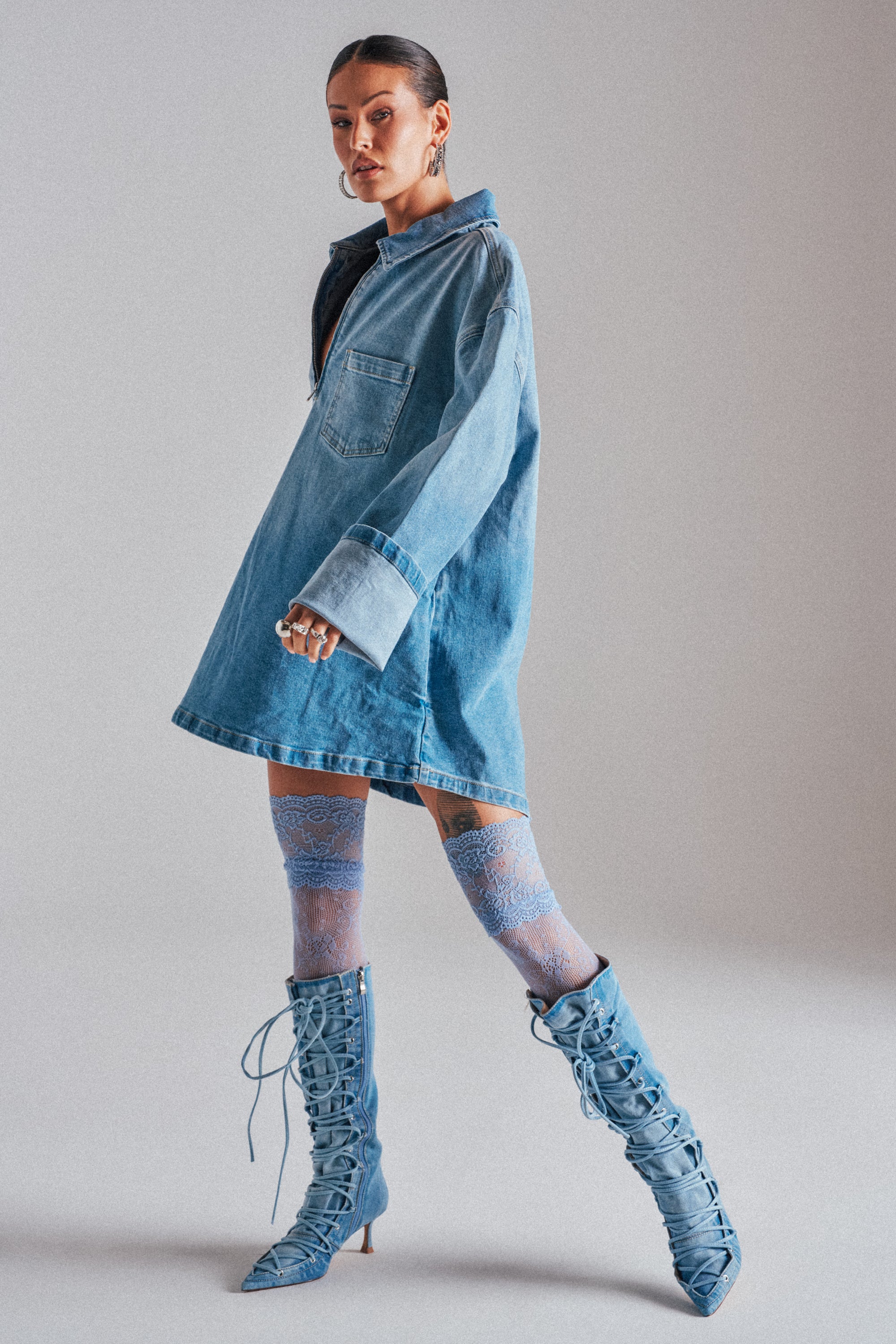 A woman models the COOL GIRL OVERSIZED OMBRE DENIM SHIRT DRESS with sheer blue lace thigh-high stockings and blue lace-up knee-high boots against a plain light background.