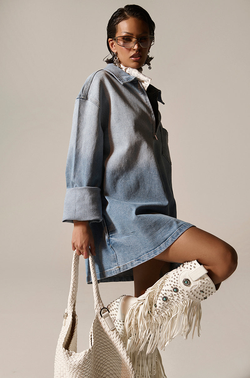 Exuding casual-cute fashion, a person poses in the COOL GIRL OVERSIZED OMBRE DENIM SHIRT DRESS, knee-length, with sunglasses and a large textured white bag. Their knee-high white boots dazzle with fringe and metallic studs, completing the effortlessly chic look.