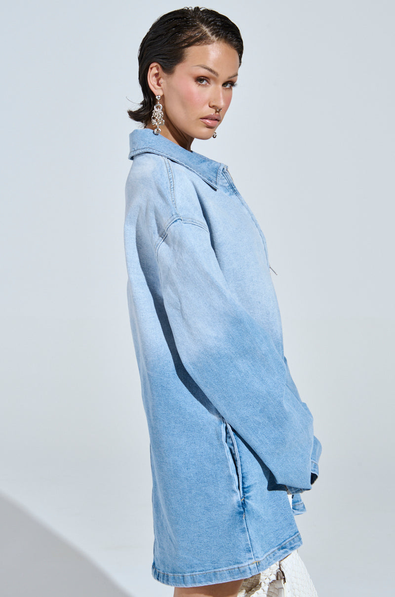 A person stands sideways in a casual-cute style wearing the COOL GIRL OVERSIZED OMBRE DENIM SHIRT DRESS. They have short, dark hair and are looking at the camera. The background is plain and light-colored, and large, ornate earrings complete their chic ensemble.