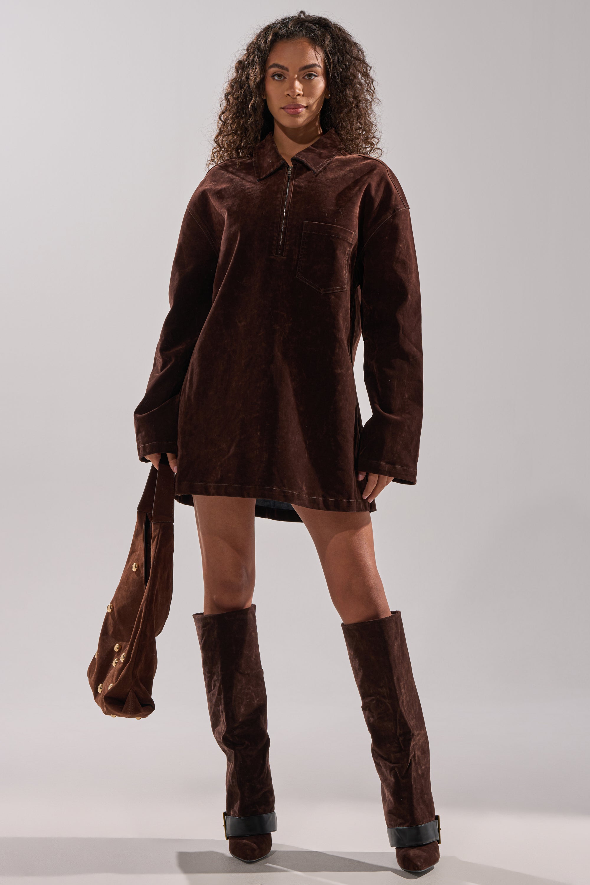 A woman with long curly hair and a neutral expression models the COOL GIRL OVERSIZED VELVET DENIM SHIRT DRESS in chocolate brown, pairing it with matching knee-high boots and a brown bag against a plain, light background.