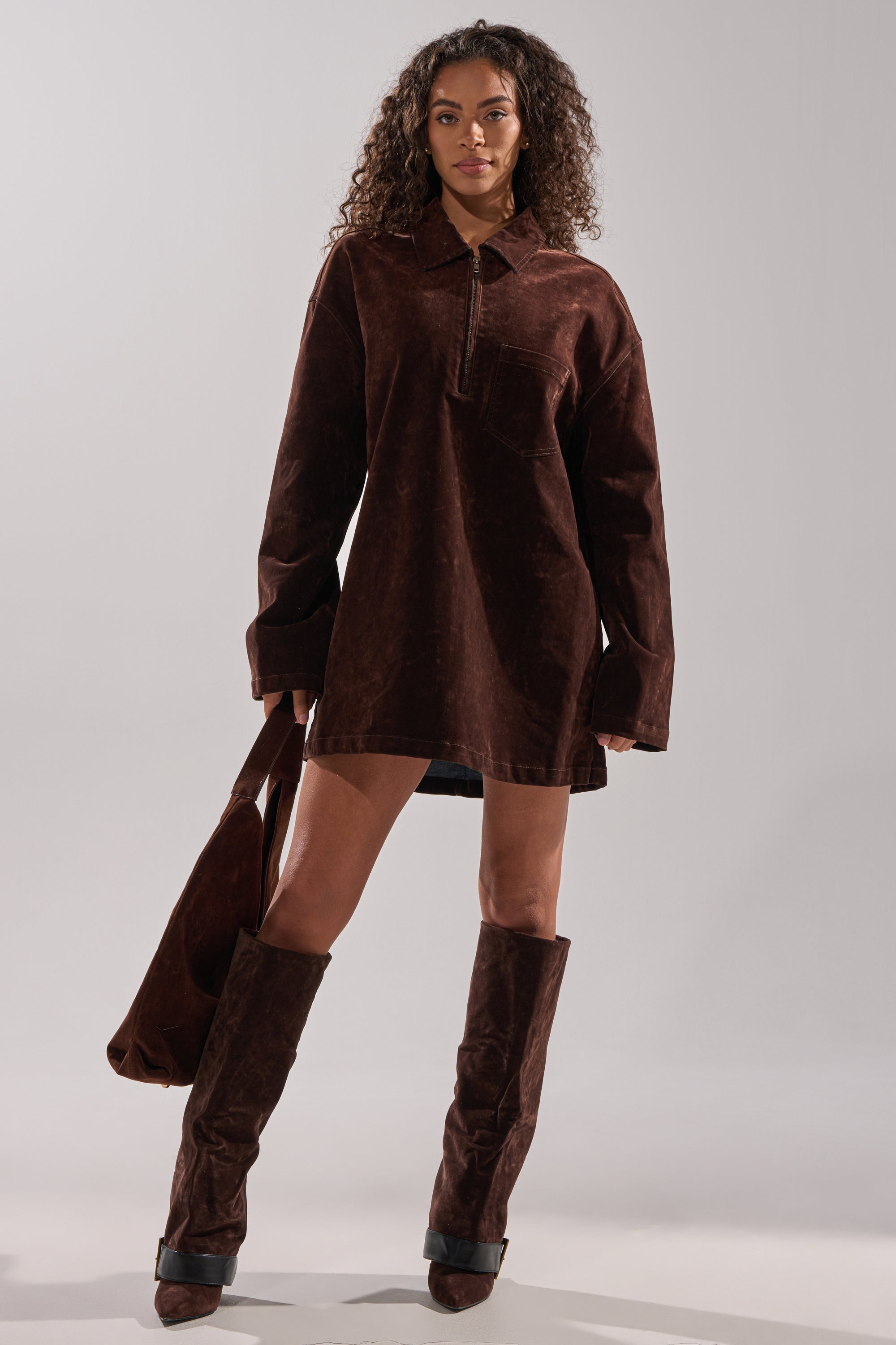 A woman exudes confidence in the COOL GIRL OVERSIZED VELVET DENIM SHIRT DRESS, paired with knee-high brown boots and a large brown bag, creating an effortlessly chic look against a light background.