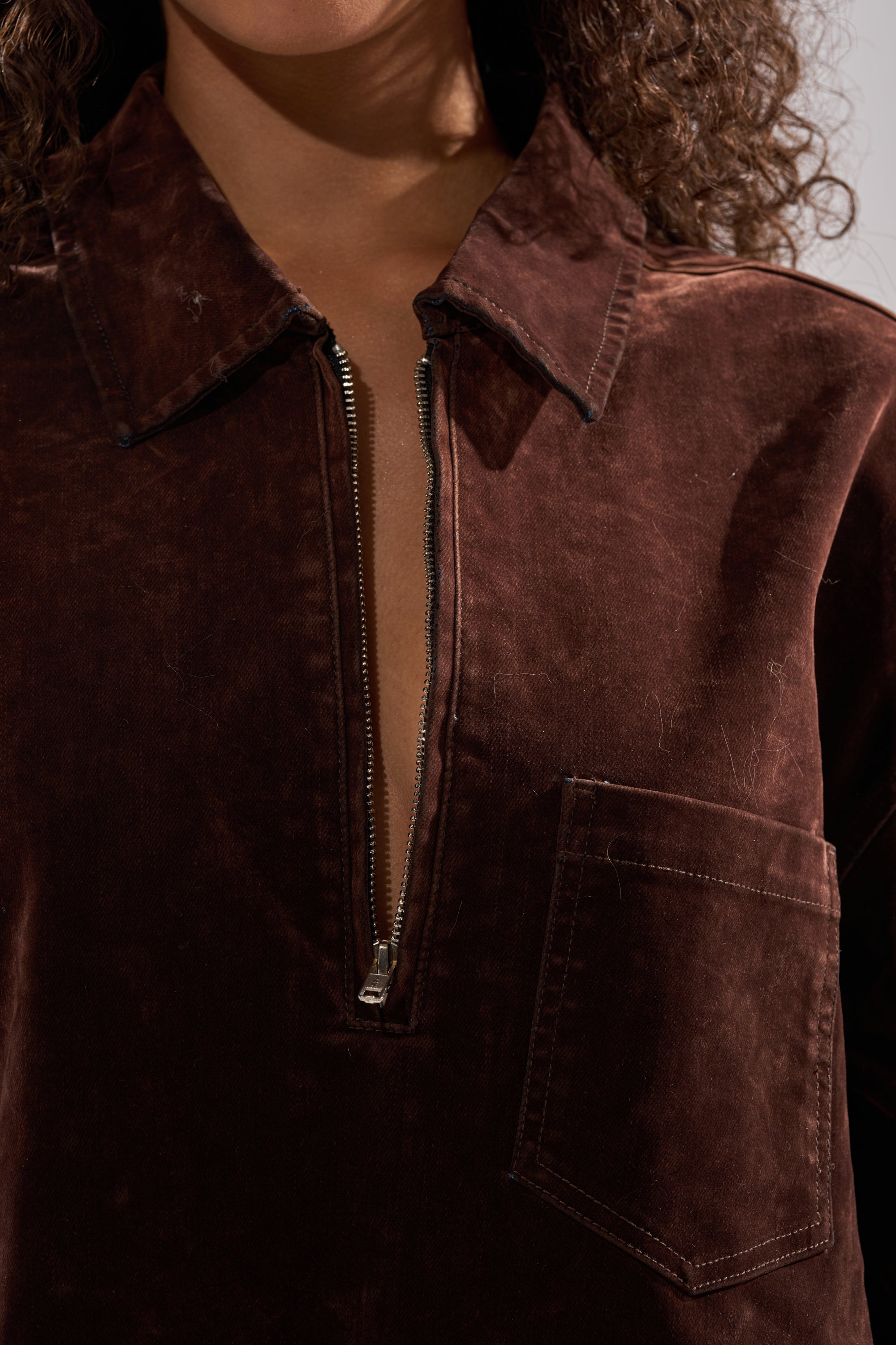 A close-up of someone wearing the COOL GIRL OVERSIZED VELVET DENIM SHIRT DRESS in chocolate brown, featuring a chest pocket and partially unzipped front. Their face is hidden, but curly hair is visible at the edges.