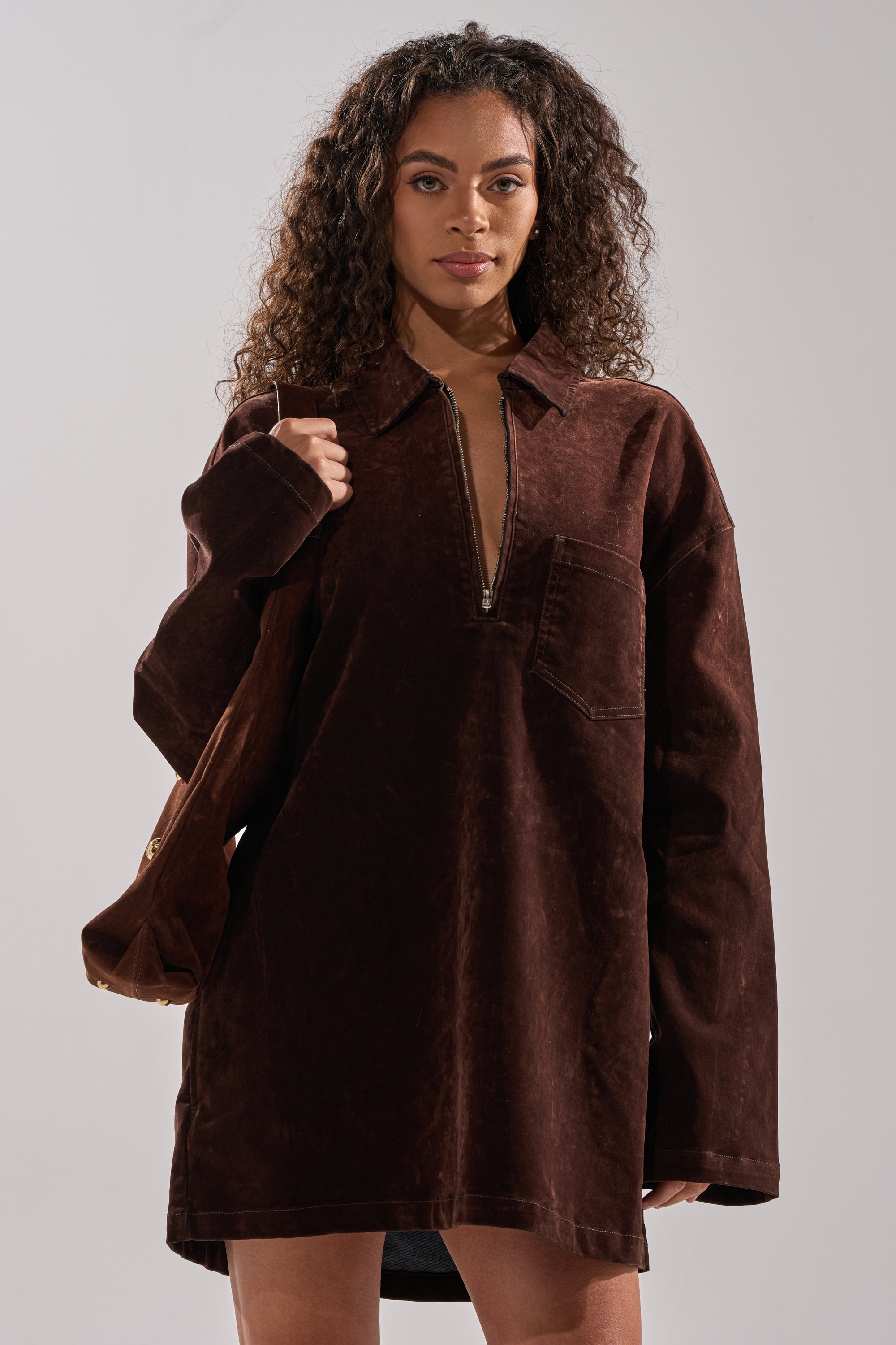 A woman with curly hair wears the COOL GIRL OVERSIZED VELVET DENIM SHIRT DRESS in chocolate brown, featuring a zipper front and chest pocket. She carries a matching brown bag over one shoulder, standing against a plain background.