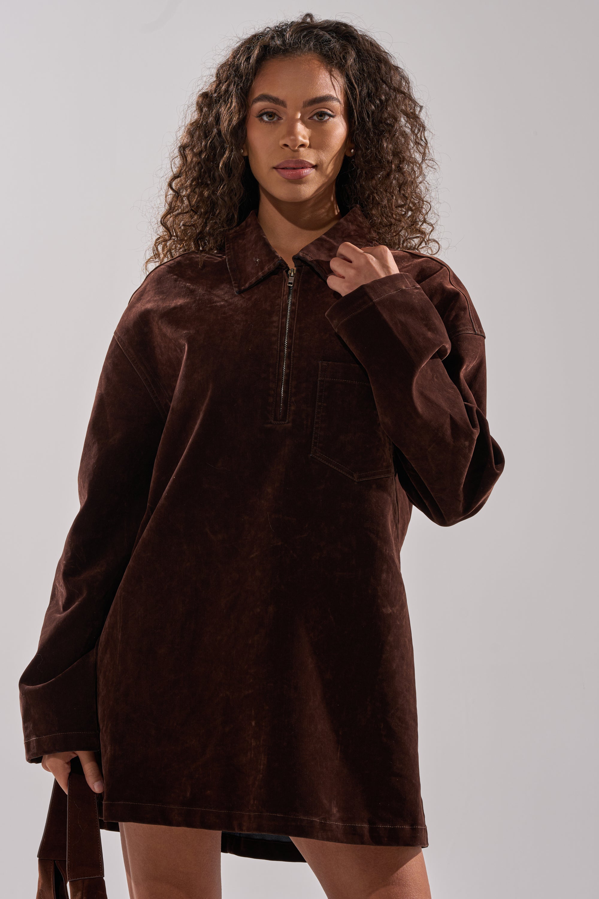 A woman with curly hair wears the COOL GIRL OVERSIZED VELVET DENIM SHIRT DRESS in chocolate brown, featuring a zipper and chest pocket, standing against a plain light background.