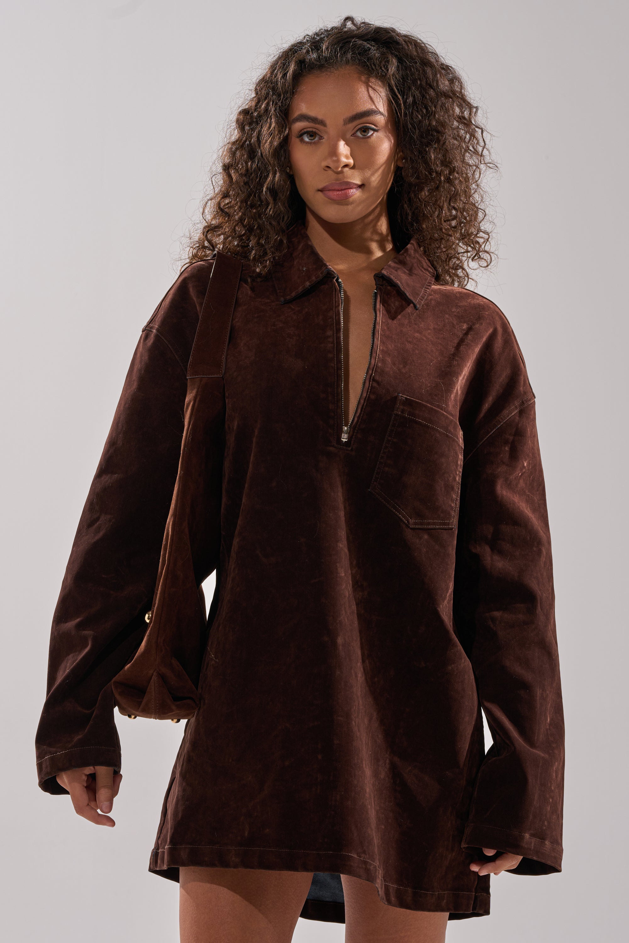 A woman with curly hair wears the COOL GIRL OVERSIZED VELVET DENIM SHIRT DRESS in chocolate brown, featuring a front zipper and chest pocket, carrying a matching shoulder bag against a plain light background.