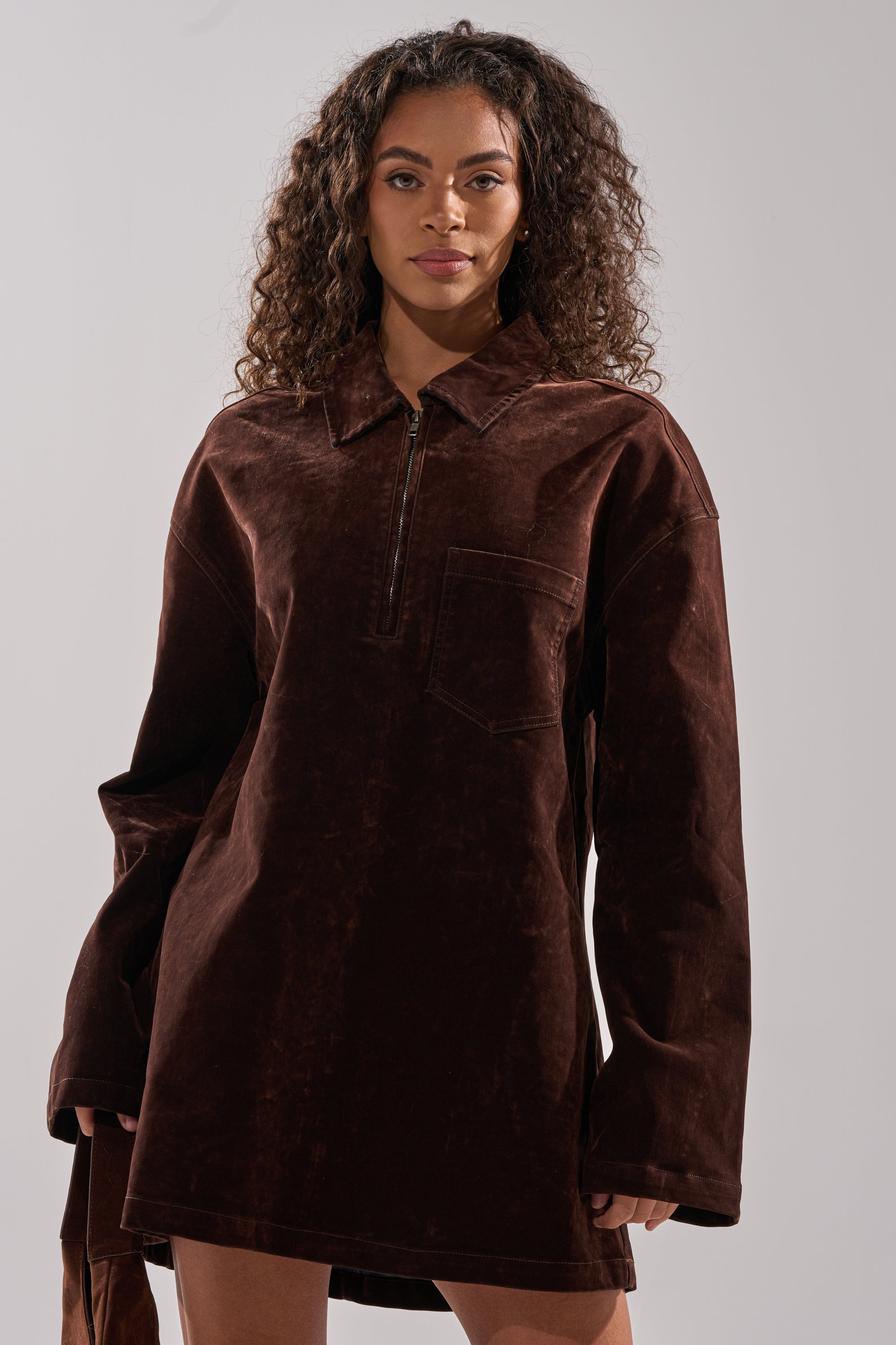 A woman with long curly hair wears the COOL GIRL OVERSIZED VELVET DENIM SHIRT DRESS with long sleeves, a collar, front zipper, and chest pocket, standing against a plain light background.