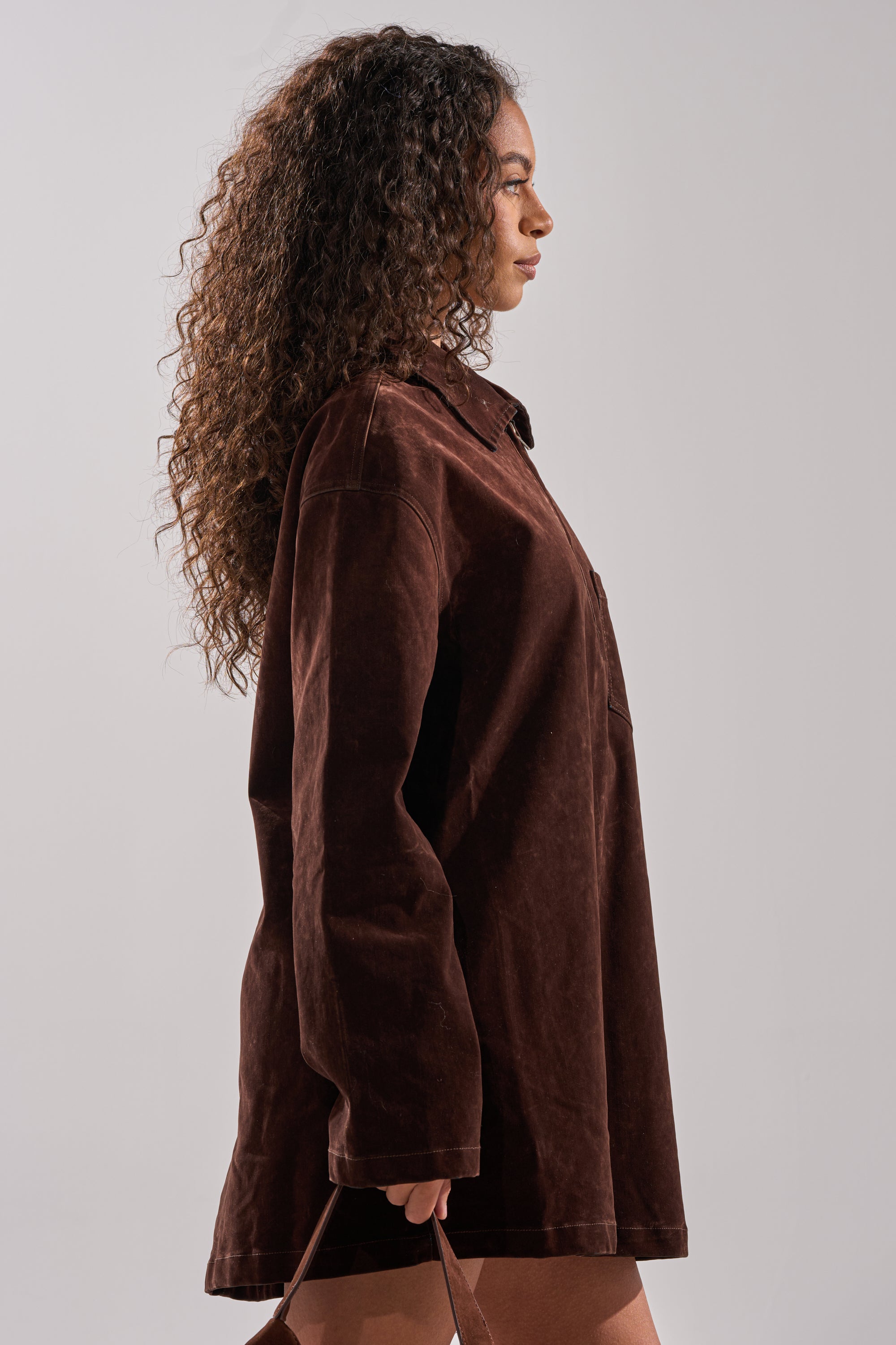 A woman with curly hair stands in profile, wearing the COOL GIRL OVERSIZED VELVET DENIM SHIRT DRESS in chocolate brown. She holds a thin strap with her right hand and looks straight ahead against a plain background.