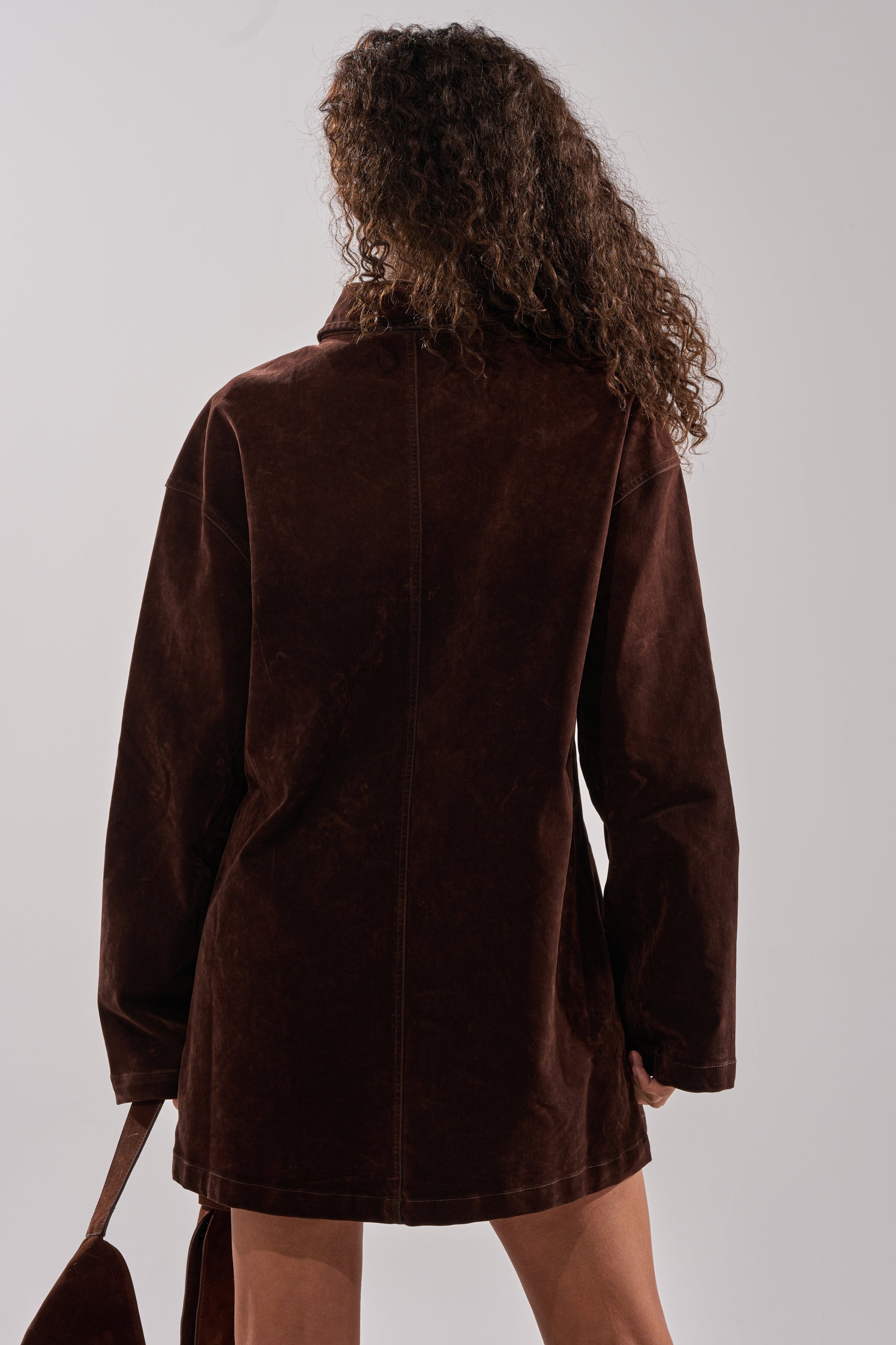 A person with curly hair stands with their back to the camera, wearing the COOL GIRL OVERSIZED VELVET DENIM SHIRT DRESS in chocolate brown and holding a matching accessory against a plain light background.